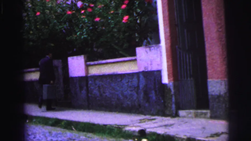 The suited man with a briefcase nears the edge of the frame as the stationary 8mm view holds on the wall, gate, and flowering shrubs.
