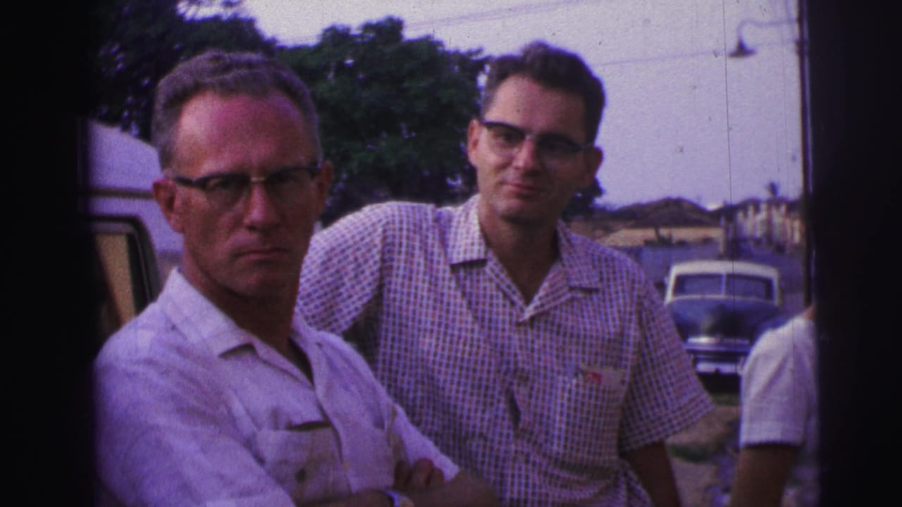 Two men in glasses stand near a parked car outdoors.