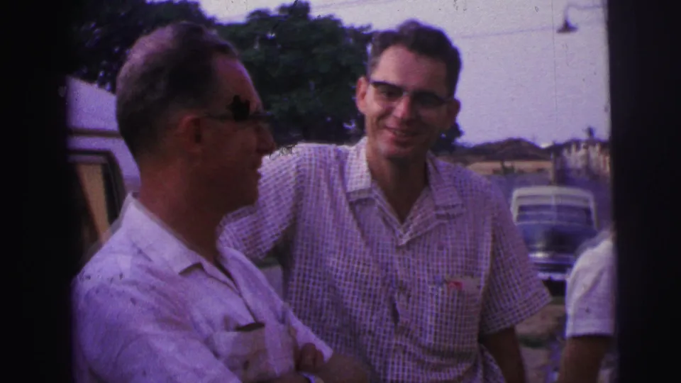 Two men beside a parked car in Brazil, one in profile and one smiling slightly toward the camera.