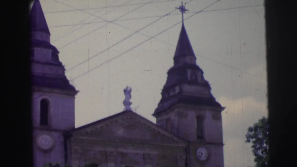 A Catholic church exterior in Brazil with twin spires, clock faces, overhead wires, and a cross above the facade.