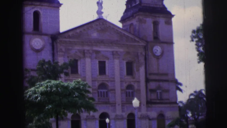 1967 Brazil 8mm view of a large church with twin towers, clock faces, arched openings, trees, and a lamppost.