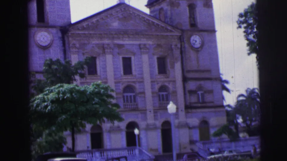1967 Brazil 8mm view of the church exterior with twin clock towers, arched doorways, railing, and tropical trees.