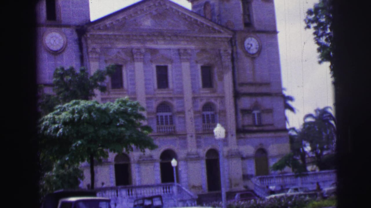 Classical church facade with clock towers, trees, and parked vehicles at the edge of the frame.