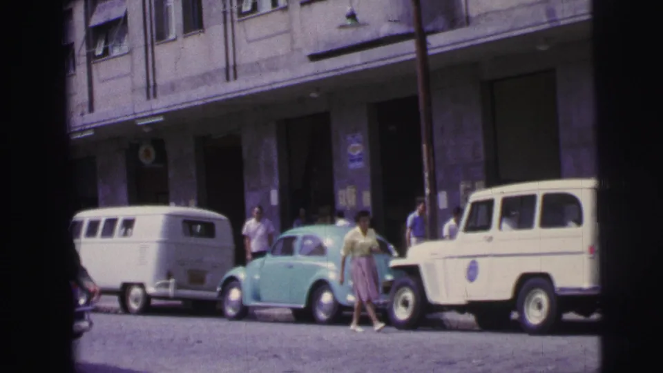 1967 Brazil street frame with a woman crossing near parked mid-century vehicles and pedestrians outside a multi-story building.