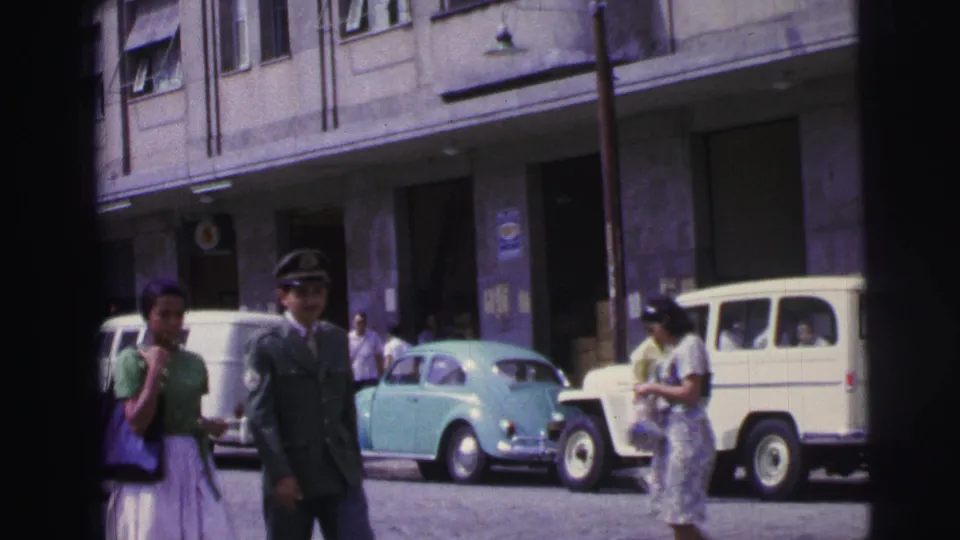 1967 Brazil street view with a man in military-style uniform, pedestrians, a light blue compact car, and a white utility vehicle.