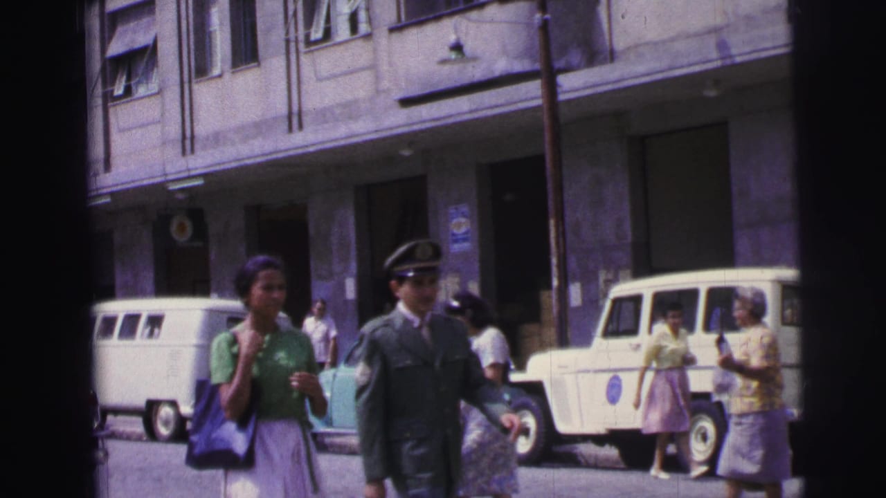 Pedestrians pass parked white vans and a pale utility vehicle outside a multi-story building.