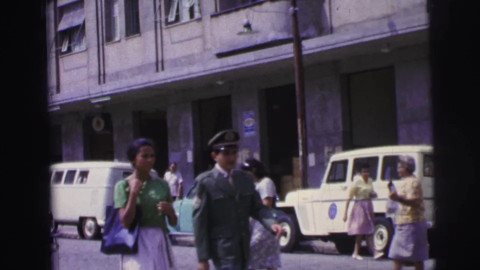 1967 Brazil street frame with a woman in green, a uniformed man, parked white vans, a pale utility vehicle, poles, doors, windows, and awnings.
