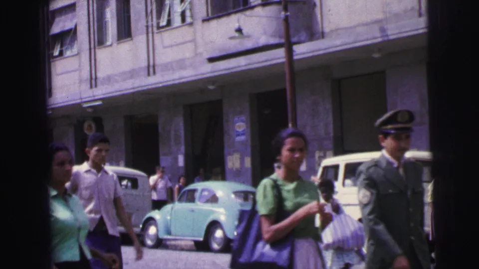 1967 Brazil urban street view with a woman carrying a bag, a uniformed man, a light blue Volkswagen Beetle, a white vehicle, and building entrances.
