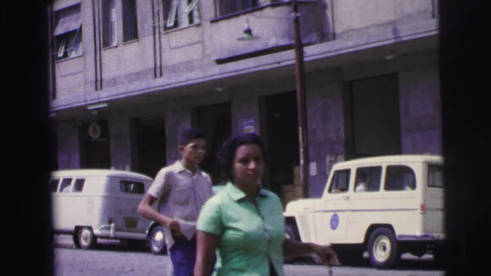 1967 Brazil sidewalk frame with pedestrians near the camera, parked white vans, a pale utility vehicle, building doors, awnings, and windows.