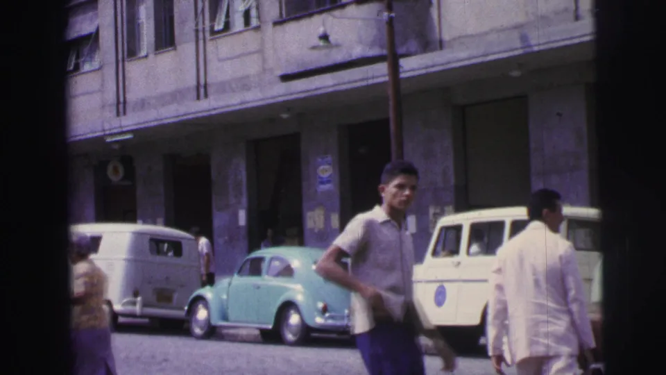 1967 Brazil street frame with a young man in the foreground, a light blue Volkswagen Beetle, white vans, a pale utility vehicle, and building frontage.