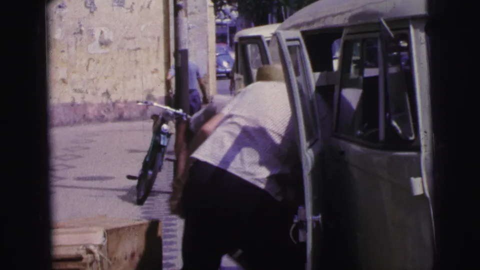 A man bends beside a light van on a Brazil street, with a leaning bicycle, weathered wall, and passing pedestrians visible.