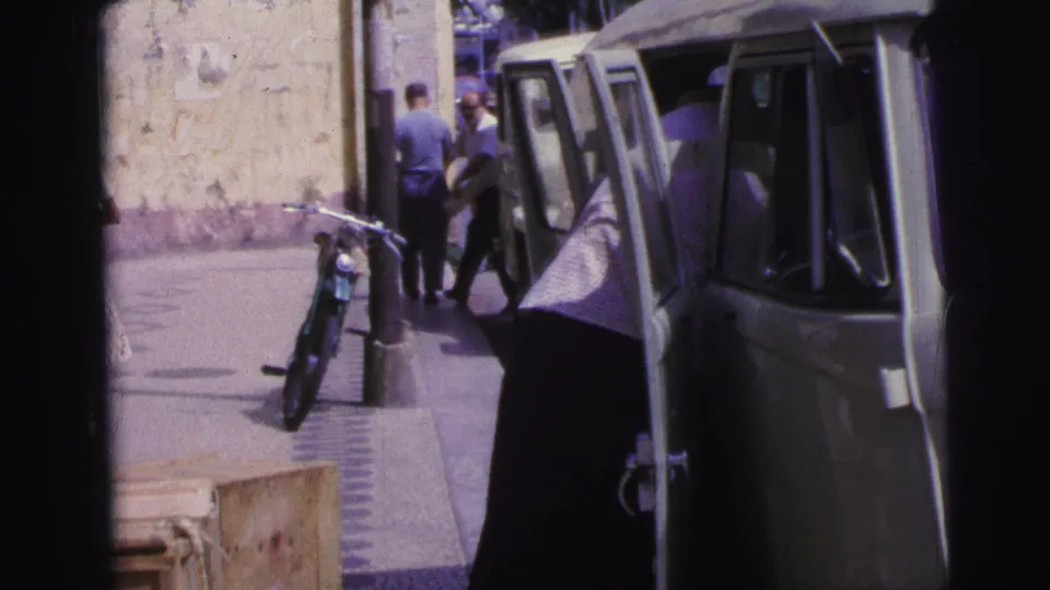 Street-level 8mm frame from Brazil showing a man leaning into a light van, pedestrians on the sidewalk, and a bicycle against a wall.