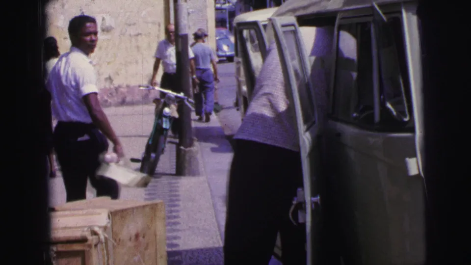 1967 Brazil street scene with a pedestrian carrying a light object, a man bent near a Kombi-style van, and a bicycle by the wall.