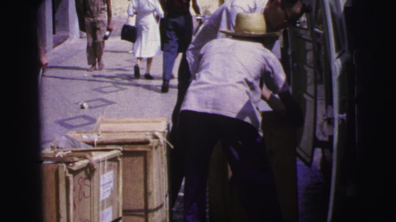 A man in a straw hat leans into a van near wooden crates as pedestrians walk past.