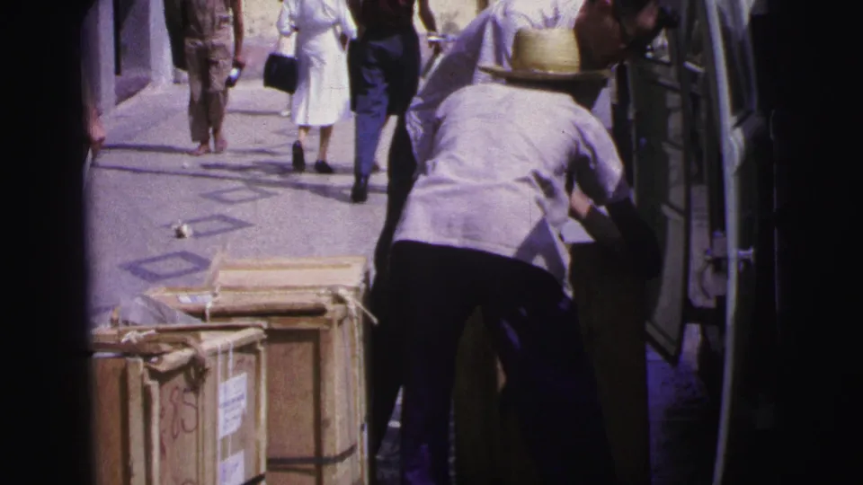 1967 Brazil street view with a straw-hatted man loading or arranging items at an open van beside wooden crates.