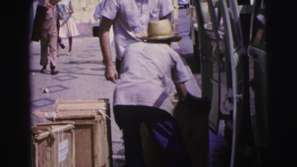 1967 Brazil home-movie frame showing a man in a straw hat bending near wooden crates beside an open van.