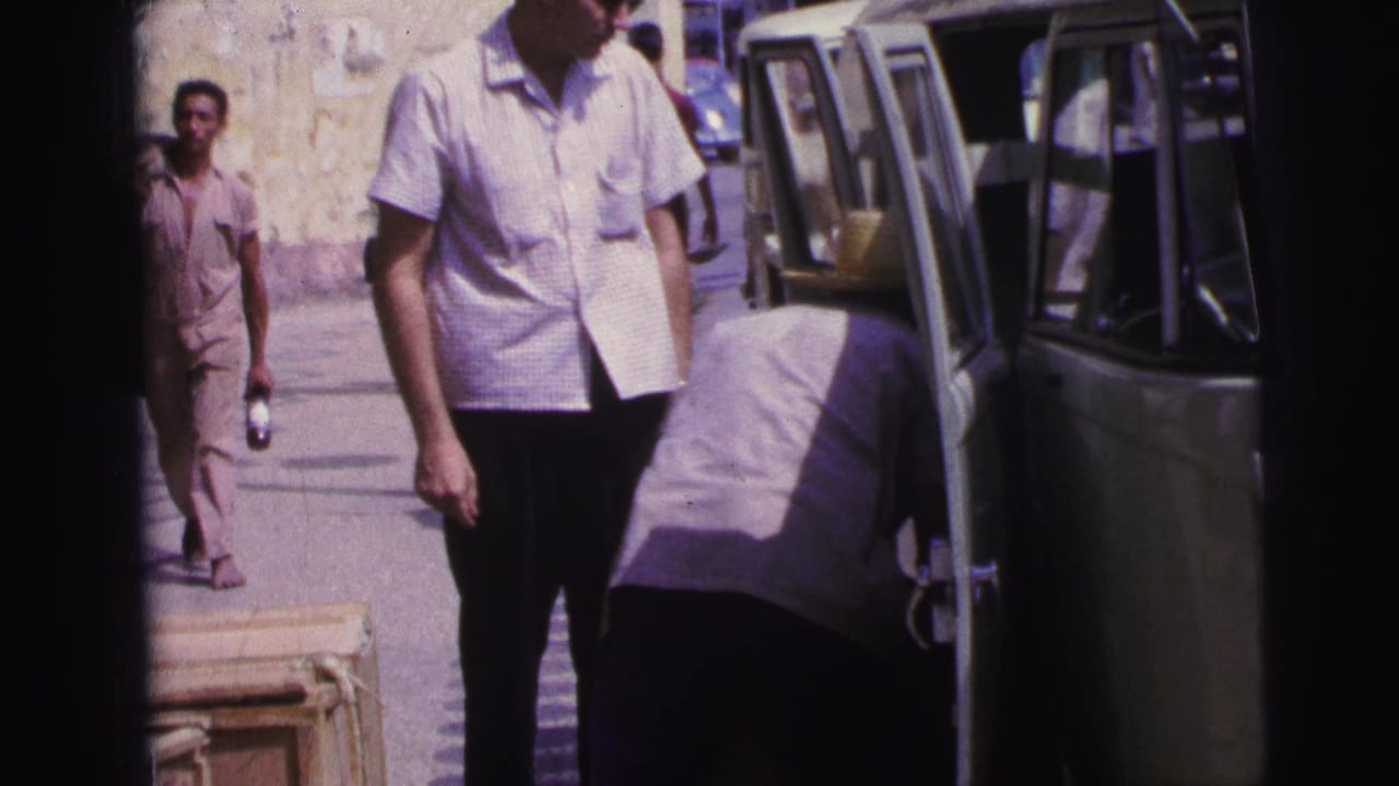 1967 Brazil home-movie frame showing men near a light green van while one person bends into the vehicle with luggage nearby.