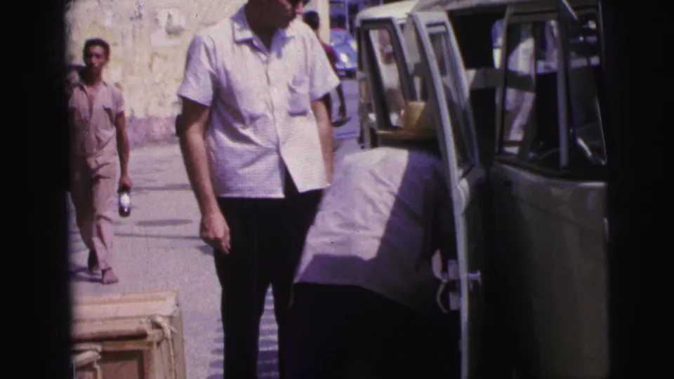 1967 Brazil home-movie frame showing men near a light green van while one person bends into the vehicle with luggage nearby.