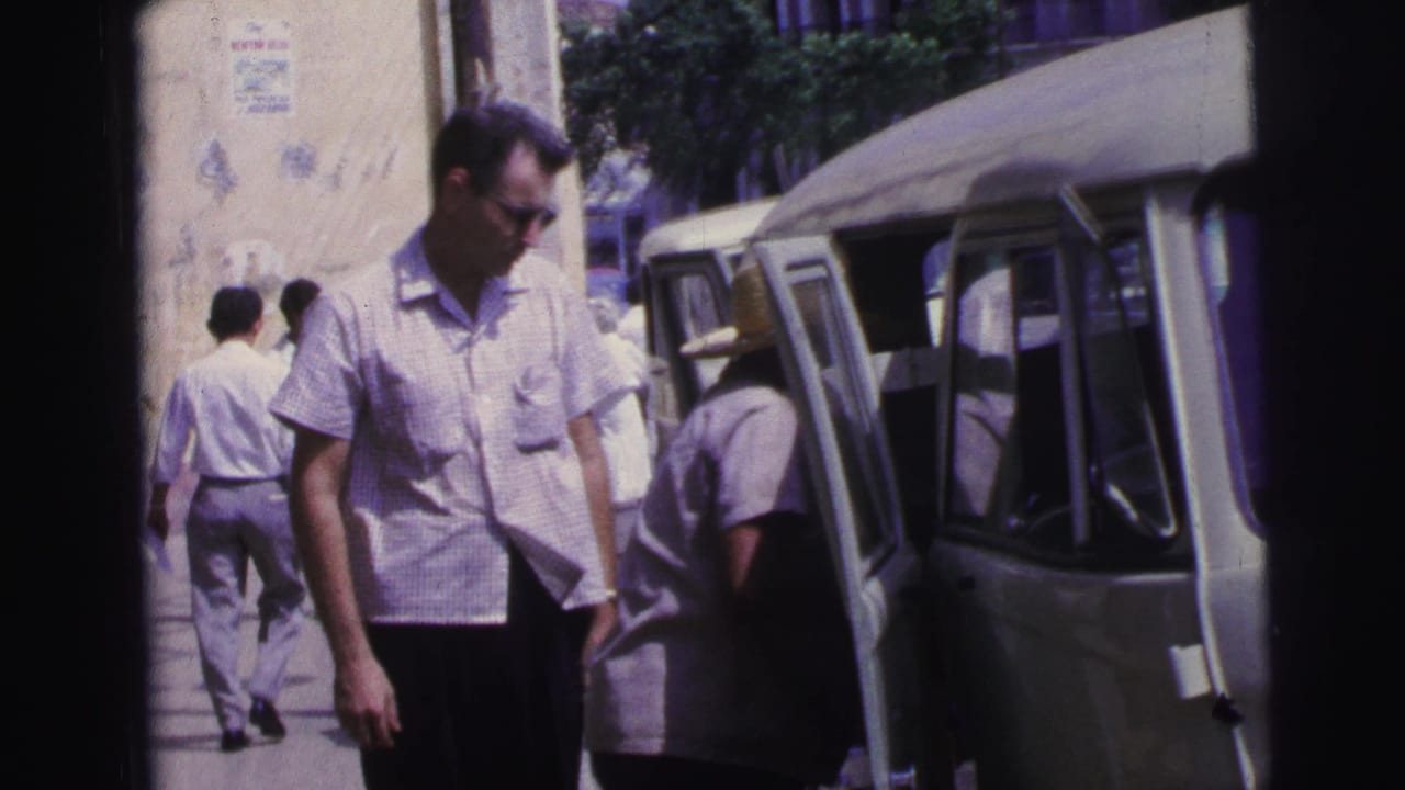 Man in sunglasses walks beside a light-colored van on a sunlit sidewalk with other adults nearby.