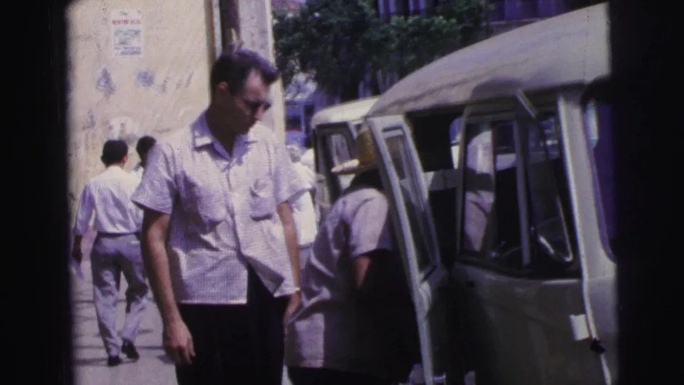 In 1967 Brazil home-movie footage, a man in a short-sleeve checked shirt and sunglasses stands near a light-colored van as adults move along the sidewalk.