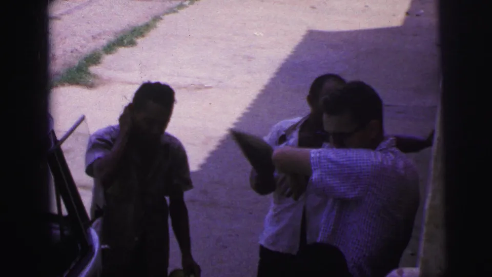 In a shaded portion of the street scene, several adults cluster by the vehicle as the man in the checked shirt leans toward another person.
