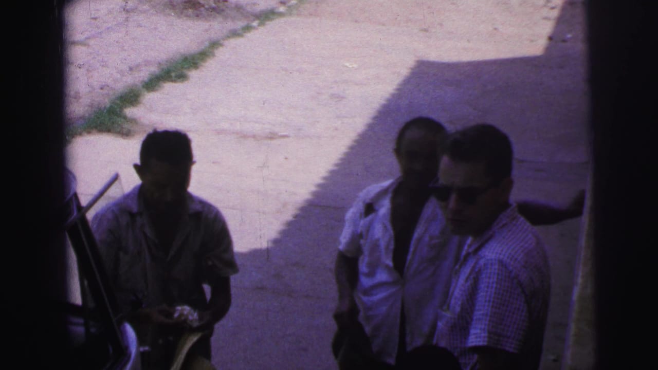 Three men stand in shade near a vehicle on a bright paved street.