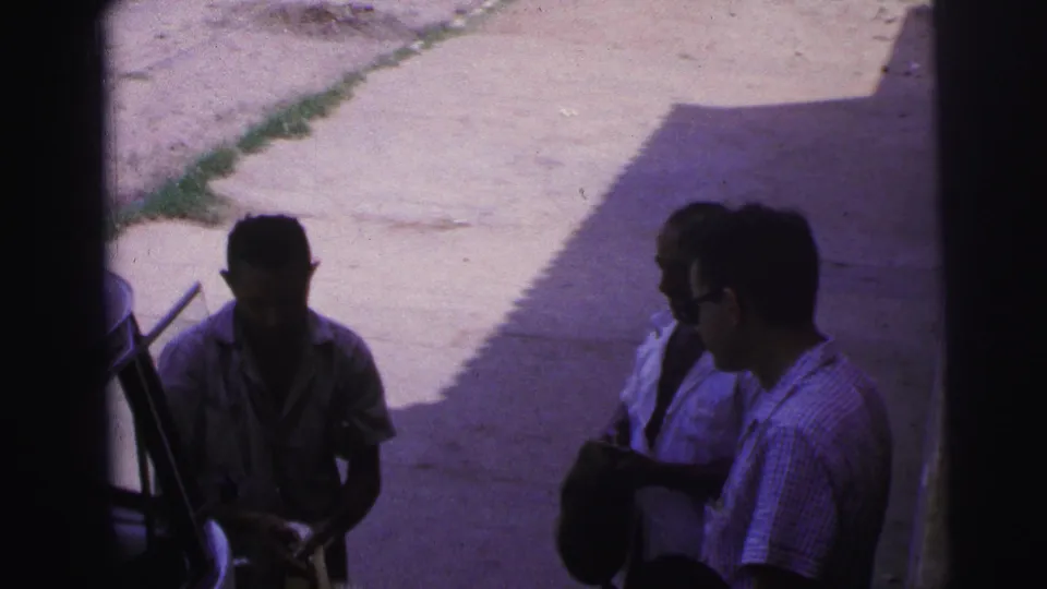 In this 1967 Brazil home-movie frame, one man in sunglasses faces another man who appears to hold a dark hat in the shade.