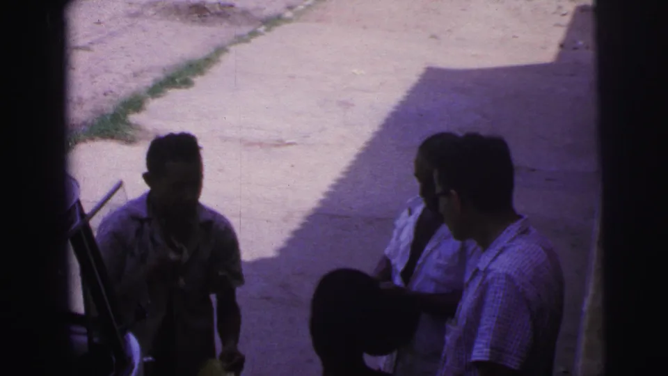 The 1967 Brazil home-movie frame shows the men still gathered in shade, with a dark hat near the foreground and sunlit pavement behind.