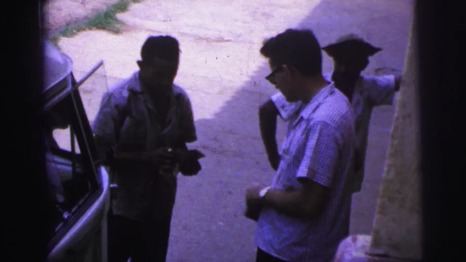 In the Brazil 1967 clip, a man in sunglasses looks down toward his hands while another man stands by the vehicle and a third remains behind.