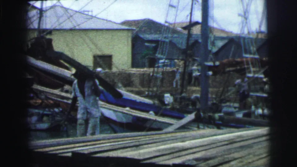 A worker in light clothing carries a long wooden piece across a Brazilian lumber or boatyard, with stacked planks and boats visible.