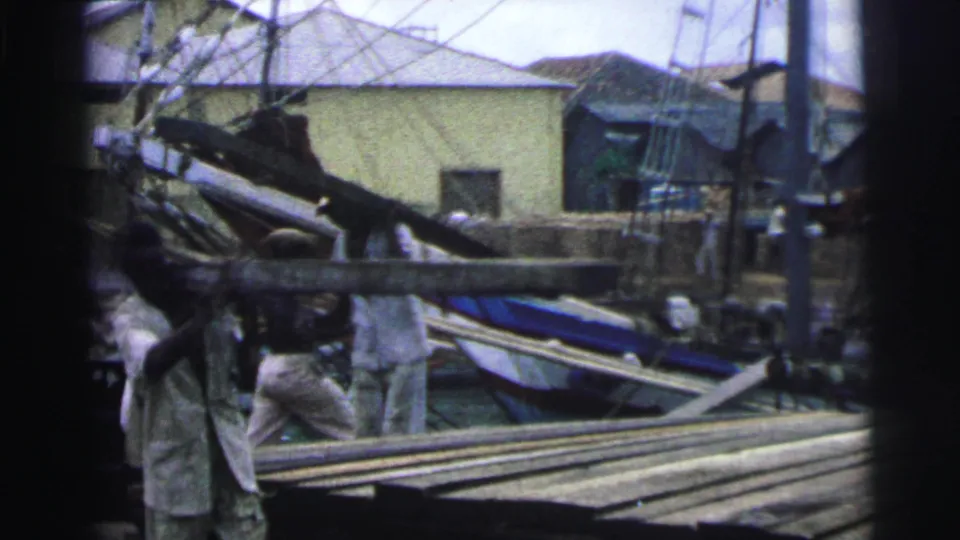 Workers continue moving heavy wooden pieces in the 1967 Brazil yard, with timber stacks, boat parts, and rigging filling the background.