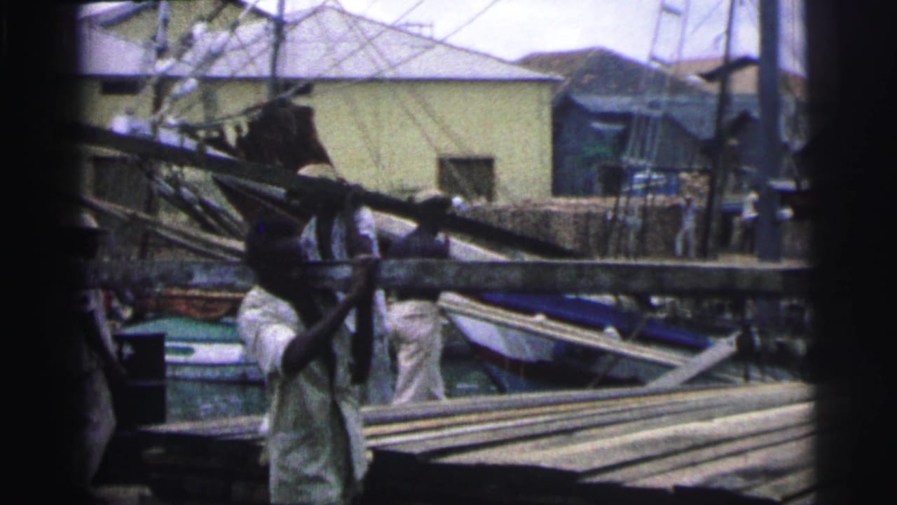 Workers carry long wooden beams near a canal with boats and pale buildings behind them.