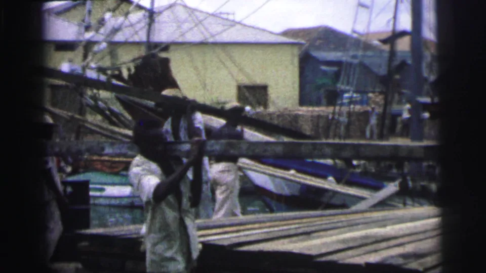 1967 Brazil 8mm frame showing men carrying long timber beside water, small boats, and low buildings in a lumber yard area.