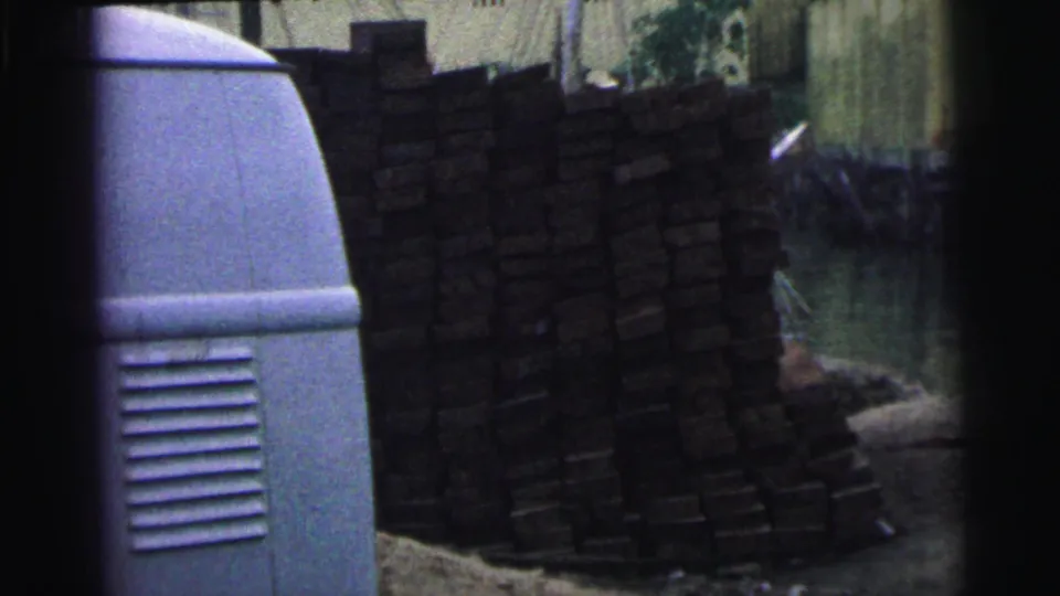 1967 Brazil 8mm frame with a tall stack of dark lumber, a pale rounded tank or vehicle side, canal water, and worksite buildings.