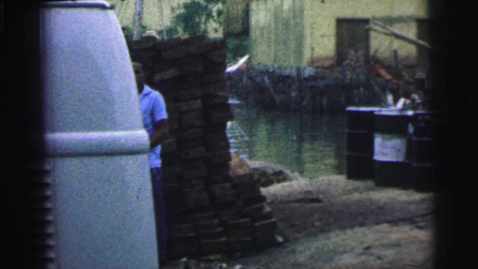 1967 Brazil 8mm frame showing stacked boards by a canal, a pale rounded tank or vehicle side, buildings, and a partially visible worker.