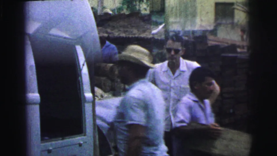 1967 Brazil 8mm frame showing several men near stacked lumber and canal water, with one man in a straw hat and another facing the camera.