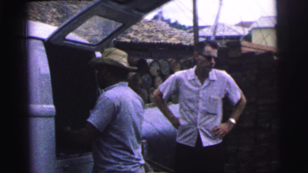 Man in straw hat stands by an open vehicle hatch while another man in sunglasses watches.
