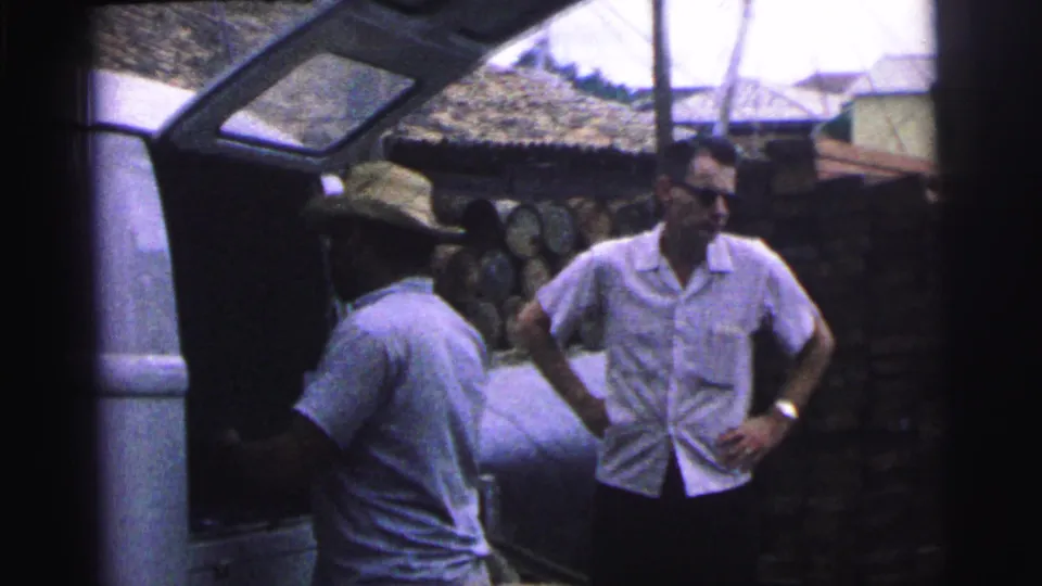 1967 Brazil 8mm frame showing a man in a straw hat near an open vehicle hatch and a second man in sunglasses standing by stacked logs.