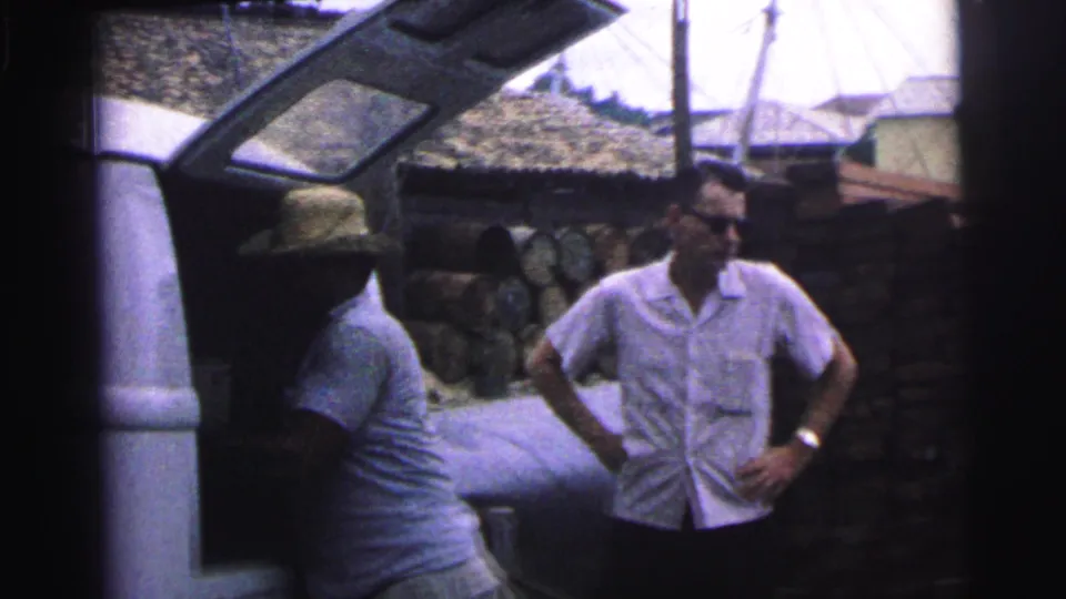 1967 Brazil home-movie frame with two men beside an open vehicle hatch, stacked logs, and simple outdoor structures behind them.