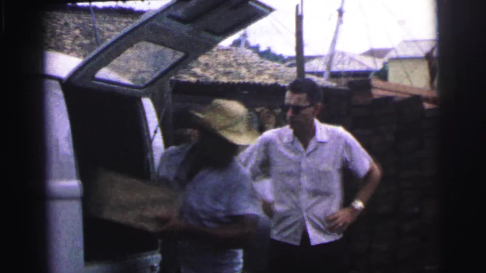 1967 Brazil frame showing a man in a straw hat working at the vehicle while a man in sunglasses stands nearby in a village-like yard.