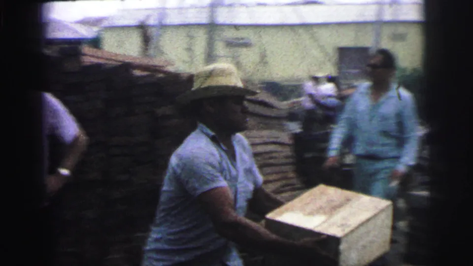 1967 Brazil 8mm frame showing a man in a straw hat carrying a wooden box across a rustic outdoor yard with another man nearby.