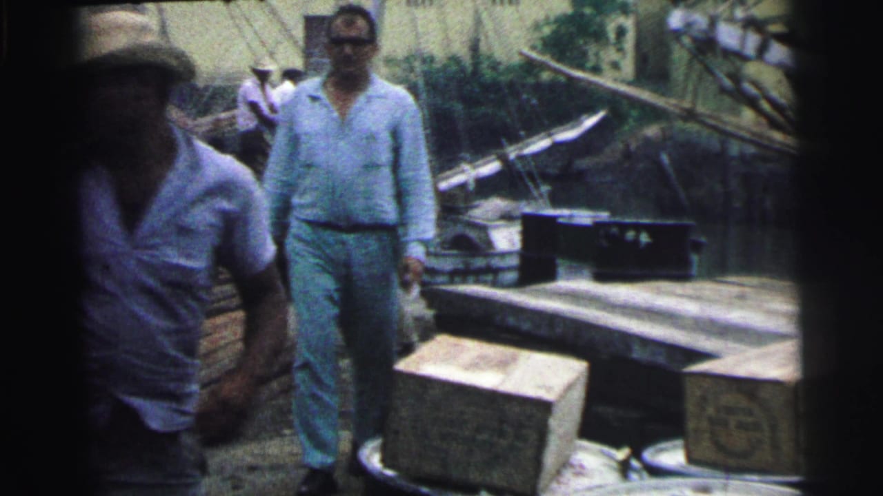 Two men stand near boxes, planks, barrels, and a boat on a dock.