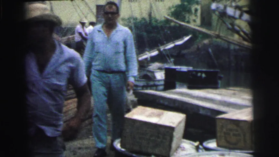 1967 Brazil 8mm frame showing men on a working dock with stacked boxes, planks, barrels, and a small boat nearby.