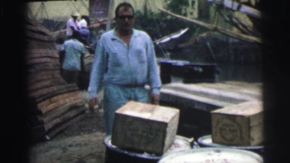 1967 Brazil dock scene with a man in light blue clothing standing beside stacked boxes, planks, barrels, and a moored boat.
