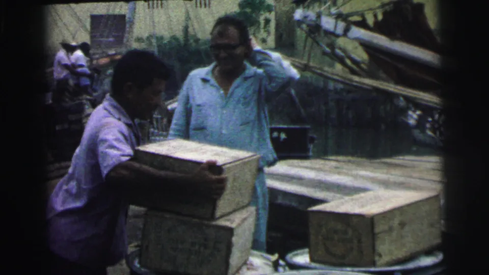 1967 Brazil 8mm frame showing a worker handling a box as another man watches near planks, barrels, and a boat.