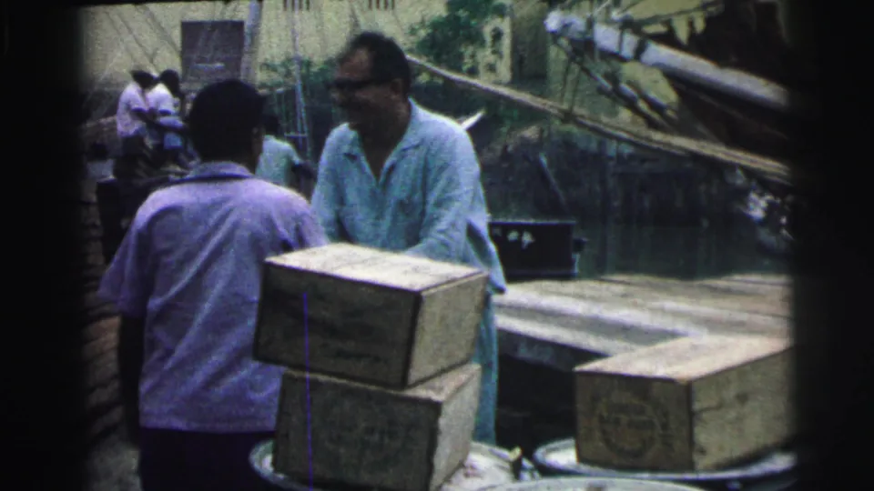 1967 Brazil dock footage with two men facing each other near boxes, barrels, planks, and a small boat in the background.