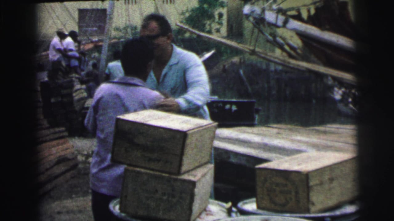 Men beside stacked wooden crates on a dock near boats and water.