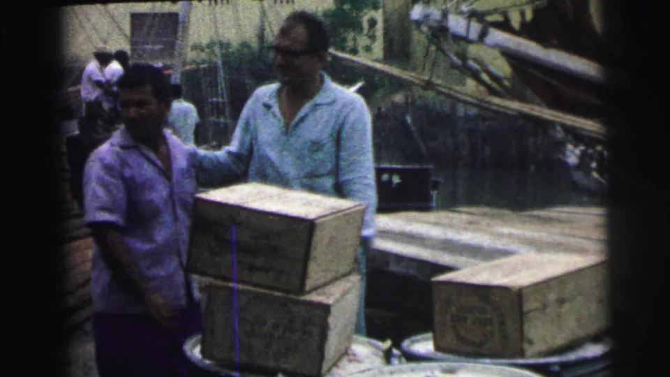 Two adult men stand by stacked wooden crates on a Brazilian dock in 1967, with boats and water visible behind them.