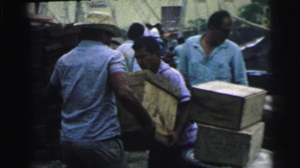 Adult dock workers in Brazil in 1967 lift or position a wooden crate near other stacked cargo on a waterfront.