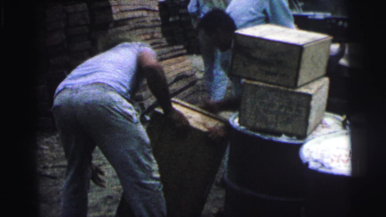 1967 Brazil home-movie frame showing workers handling wooden crates beside barrels on a dock or quayside.